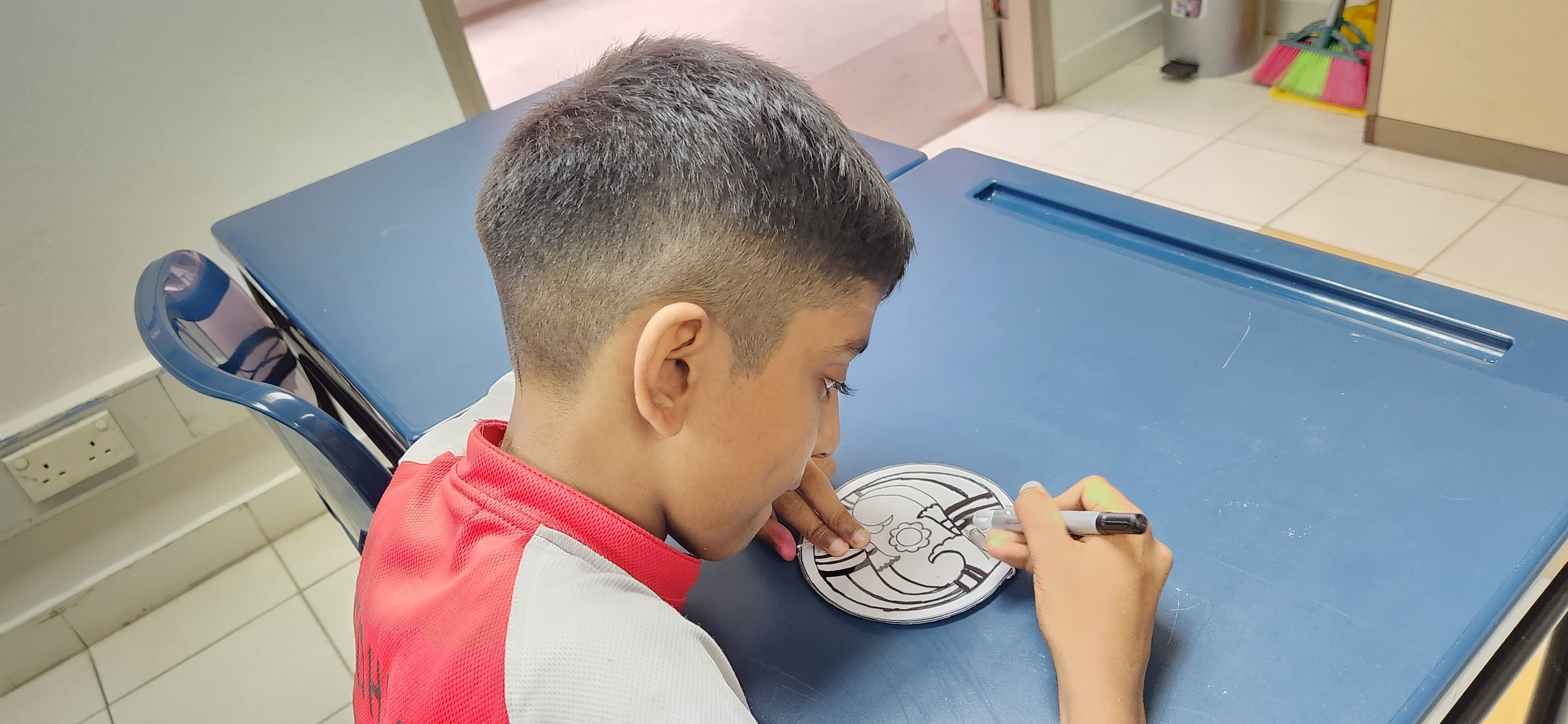 A young boy leaning over a blue desk, drawing on a small round disc with a black marker.